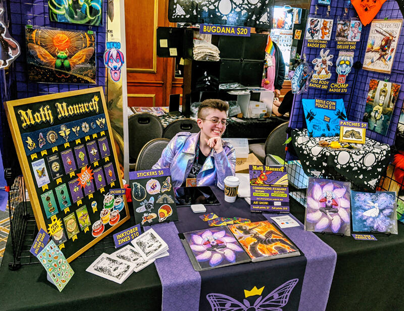 The artist seated behind their fully decorated vendor table.